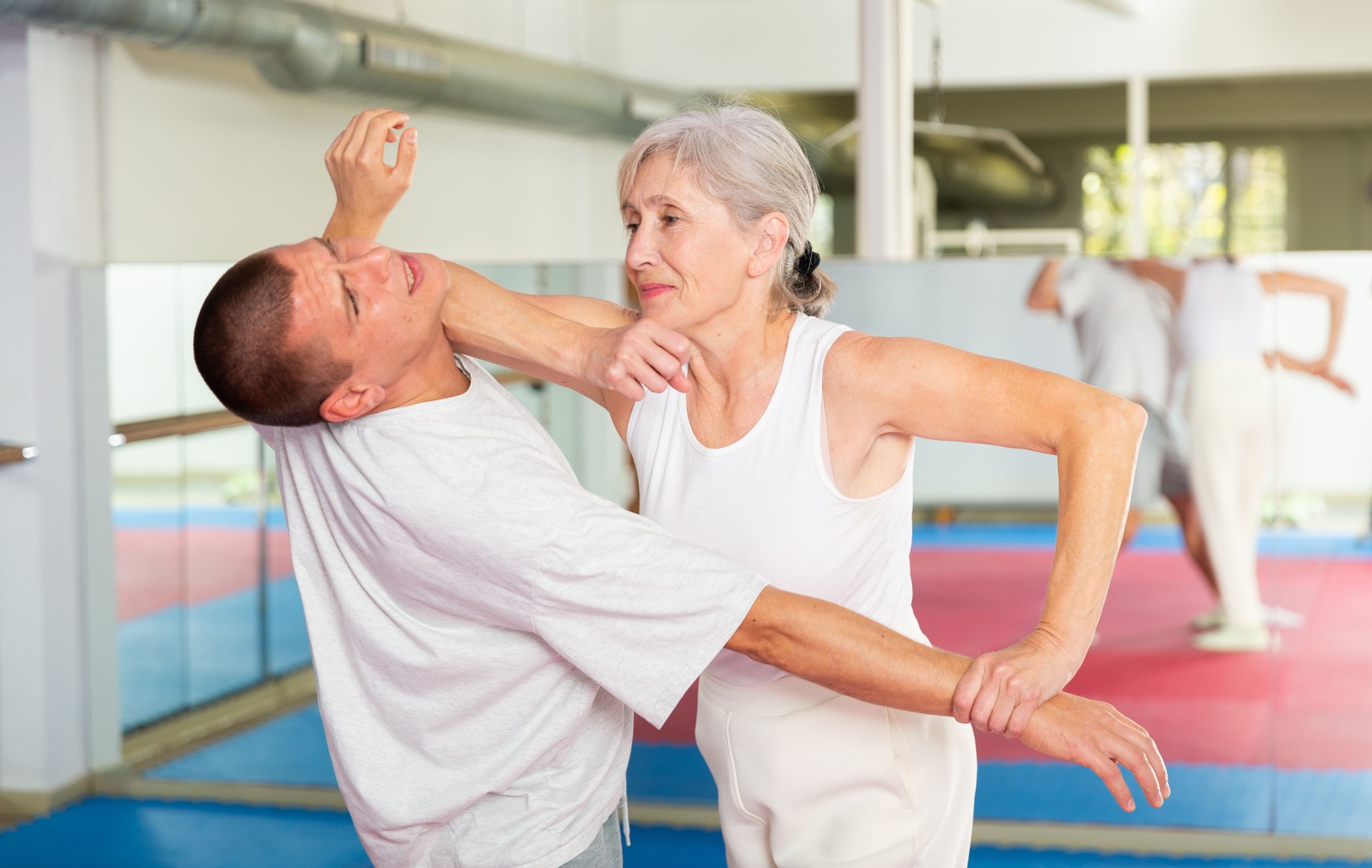 Aged woman practicing elbow blow with wristlock to opponent in gym